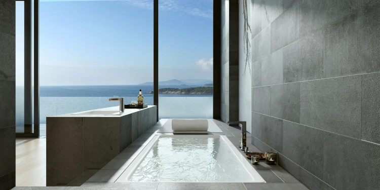 Salle de bain contemporaine avec baignoire encastrée, mur en pierre grise et grandes baies vitrées sur la mer.