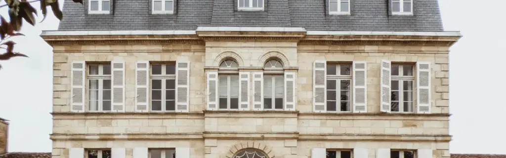 Façade de maison de maître en pierre de taille avec toiture en ardoise, lucarnes, volets blancs et entrée cintrée.