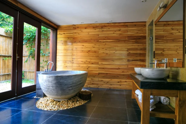 Salle de bain style spa avec baignoire en pierre, mur en bois, sol sombre, vasques et grande baie vitrée sur jardin.
