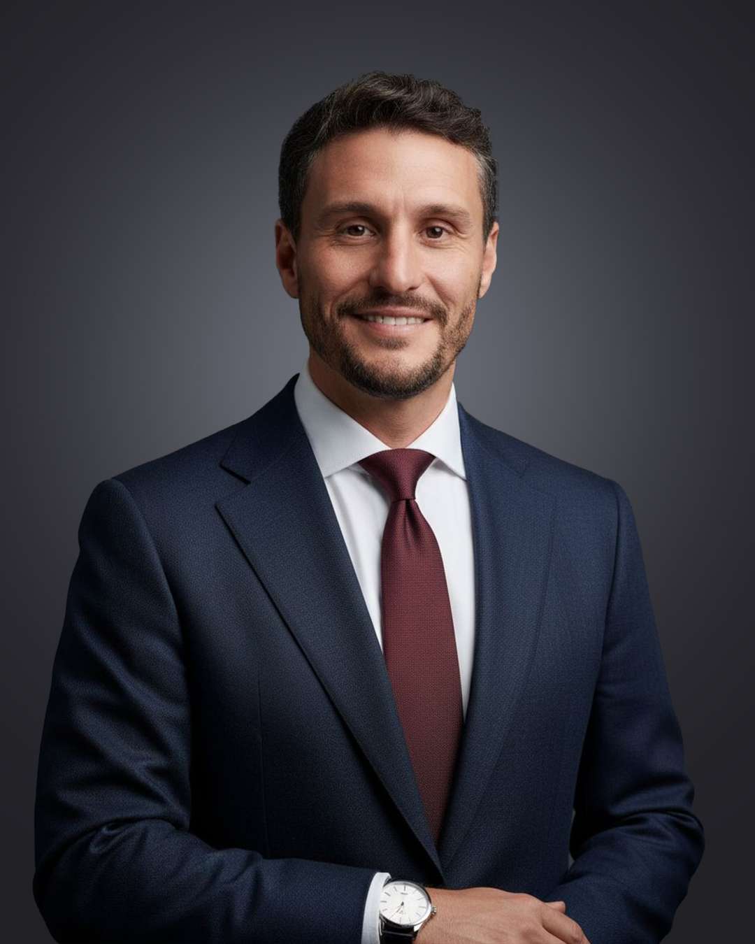Portrait studio d’un homme en costume bleu et cravate bordeaux, souriant, sur fond gris.