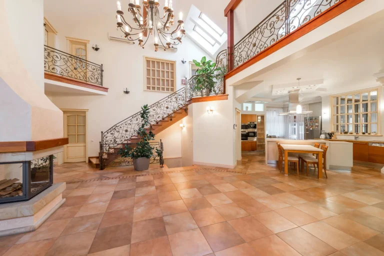 Grande cuisine ouverte de villa avec sol en terre cuite, escalier en fer forgé, cheminée et double hauteur sous verrière.