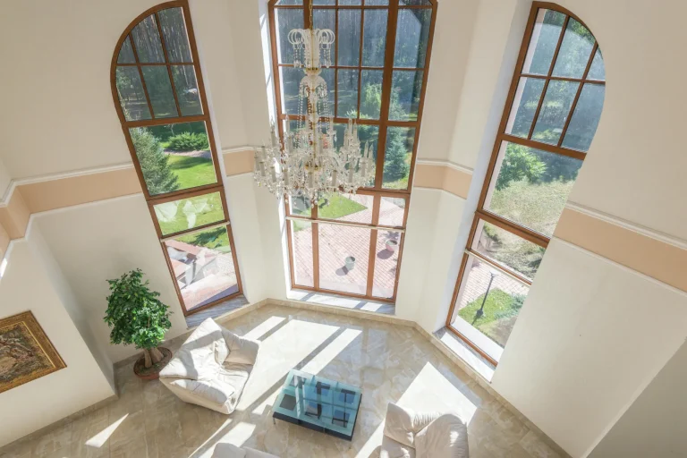 Intérieur maison de maître avec salon double hauteur, grandes baies cintrées, lustre en cristal et vue sur jardin verdoyant.